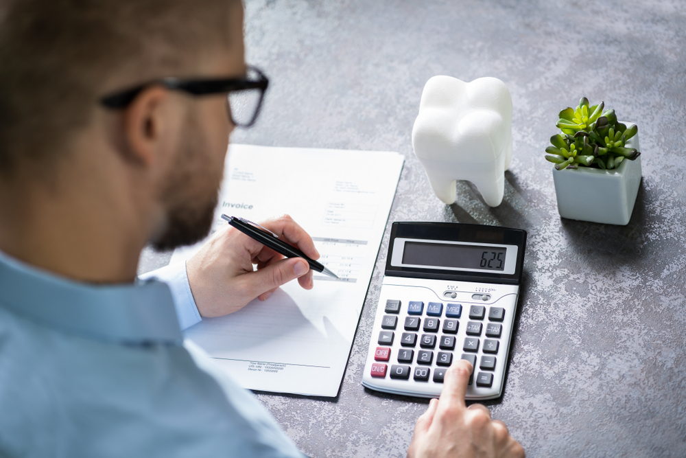 Close-up Of White Tooth Over Desk In Front Of Man Calculating Bill With Calculator